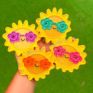 May include: Four yellow sun-shaped decorations with smiling faces and sunglasses in various colors: pink, green, and orange. The suns have a textured, felt-like appearance and are held against a green background.