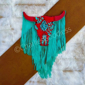 May include: Red and turquoise bull skull with turquoise fringe hanging from the bottom. The skull is decorated with turquoise and copper accents. The background is a white faux fur.