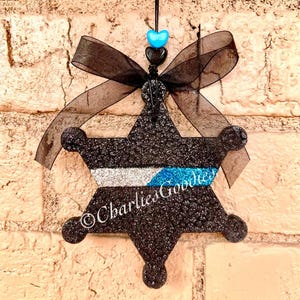May include: Black glitter police badge ornament with a thin blue line and silver glitter. The ornament is decorated with a black ribbon and blue and black heart beads.