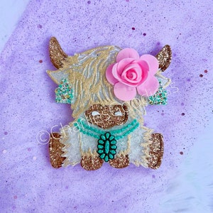 May include: A white and gold glitter Highland cow with a pink flower and turquoise necklace. The cow is sitting down and has a brown nose.