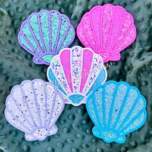 May include: Five glitter seashell ornaments in different colors: purple, pink, white, light blue, and blue. The seashells are on a green textured surface.