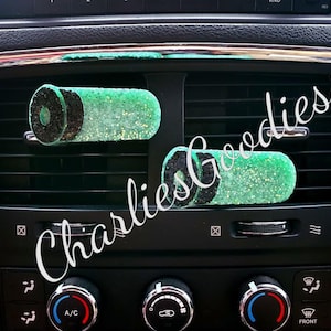 May include: Two green and black car air fresheners shaped like bullet casings. The air fresheners are hanging from the air vent in a car dashboard.