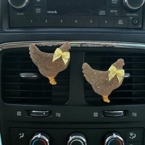 May include: Two brown glitter chicken-shaped car air fresheners with yellow bows hanging from the air vents in a car.