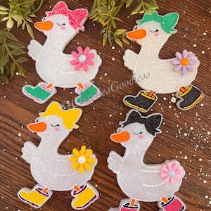 May include: Four glittery white goose ornaments with colorful bows, flowers, and boots. Each goose has an orange beak and is wearing boots in different colors: green, black, yellow, and pink. The ornaments are on a brown surface.