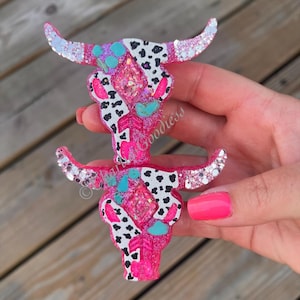 May include: A pair of decorative bull skull ornaments. The skulls are primarily hot pink with black and white cow print accents. They feature glitter horns and turquoise and pink embellishments. The word "Goodiess" is visible on the bottom skull.