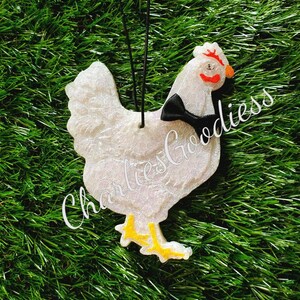 May include: White chicken-shaped ornament with a black bow tie, orange beak, and red comb. The ornament has yellow feet and the text "Charlies Goodiess" in a cursive font. The ornament hangs from a black string and is set against a green background.