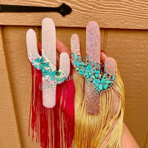 May include: Two cactus-shaped ornaments. One is white with turquoise accents and red fringe, the other is rose gold with turquoise accents and gold fringe. Both have glitter details.