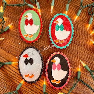 May include: Four oval-shaped Christmas ornaments featuring white geese with bow ties. Each ornament has a different background color: green, red, white, and black. The ornaments are surrounded by a string of Christmas lights.