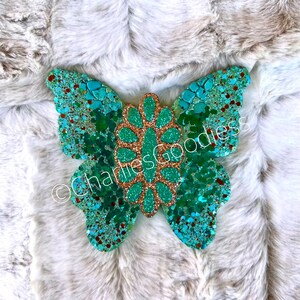 May include: A turquoise and gold glitter butterfly with a floral design in the center. The butterfly is on a white fuzzy background.