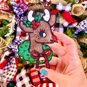 May include: A brown reindeer with a green wreath around its neck. The wreath is decorated with red berries and a red bow with a gold bell. The reindeer has a white face with brown eyes and a brown nose.