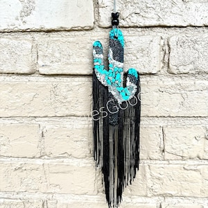 May include: A black and turquoise cactus wall hanging with black fringe. The cactus is made of mosaic tiles and has a white outline.
