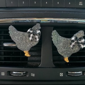 May include: Two silver glitter chicken car air fresheners with black and white checkered bows hanging from the air vents of a car.