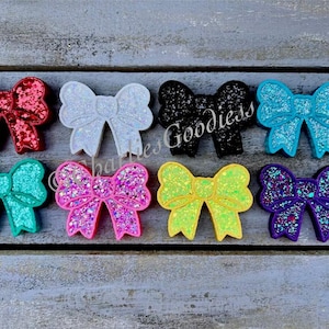 May include: Ten different colors of glitter bows. The bows are all the same size and shape, and they are arranged in a row on a wooden surface. The colors of the bows are red, white, black, turquoise, teal, pink, yellow, purple, and two shades of blue.