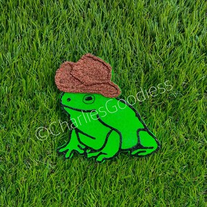 May include: A green frog wearing a brown cowboy hat. The frog is sitting on a green grassy surface.
