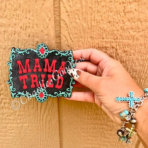 May include: A black sign with the words "MAMA TRIED" in red lettering. The sign has a turquoise border with red accents. The sign is held in front of a wooden background. A bracelet with turquoise beads and cross charms is also visible.