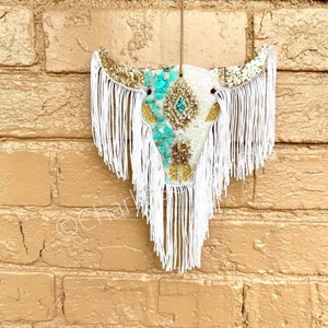 May include: White cow skull with turquoise and gold accents, decorated with white fringe.