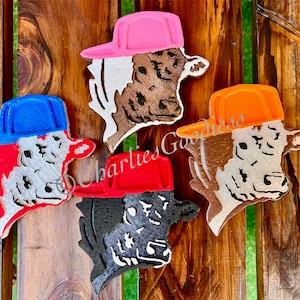 May include: Four wooden cow head ornaments with different colored baseball caps. The caps are pink, blue, orange, and red. The cow heads are painted white with black markings.