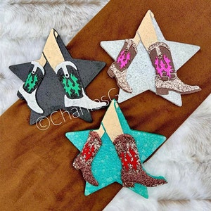 May include: Three wooden star-shaped ornaments with a pair of legs and boots on each. The boots are decorated with glitter and have a western design. The stars are painted in black, white, and turquoise.