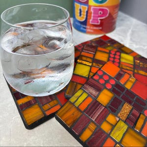 Op de afbeelding: Een glas water met ijsblokjes staat op een mozaïeken onderzetter. De onderzetter heeft een geometrisch ontwerp in rood, oranje en geel. Op de achtergrond is een blikje met het woord "UP" te zien.