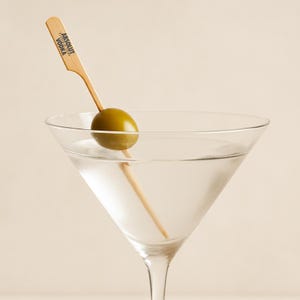May include: A clear martini glass filled with a clear liquid, with a green olive on a wooden skewer. The skewer has the text "Absolut Vodka" printed on it. The glass is set against a light beige background.