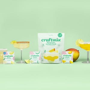 May include: A variety pack of margarita mix packets with a green and white design. The packets are arranged on a green surface with a glass of margarita, limes, and a mango.