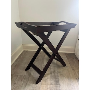 May include: A dark brown wooden folding tray table with a rectangular tray top and cut-out handles. The legs are in an X-shape, and the table is set on a light-colored wooden floor.
