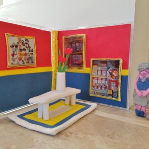 May include: A miniature diorama of a room with red and blue walls, a white table, a yellow and blue rug, and a vase with red flowers. Two figures stand in the doorway, one wearing a blue hat and the other wearing a grey hat.