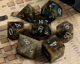 May include: A set of eight polyhedral dice made of green labradorite stone with silver numbers. The dice are on a brown and white background with a grid pattern.