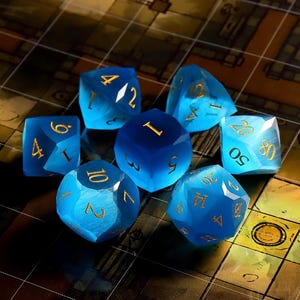 May include: A set of seven translucent blue polyhedral dice with a cat's eye effect and engraved gold numbers. The collection includes various shapes such as a d20, d12, d10, d8, d6, and d4, all featuring smooth, faceted surfaces and metallic gold ink.