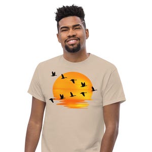May include: A beige short-sleeved t-shirt featuring a graphic print of a flock of black birds flying in front of a large, bright orange sunset. The sun is positioned centrally on the chest, with its reflection visible at the bottom of the design.