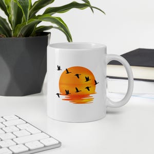 May include: A white ceramic mug featuring a graphic print of a vibrant orange and yellow sunset over water with a flock of black birds flying across the sky. The design includes a reflection of the sun on the water surface and a curved handle.