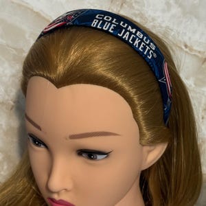 May include: A navy blue headband with the text "COLUMBUS BLUE JACKETS" in white. The headband features a repeating pattern of the team's logo and colors. The headband is displayed on a mannequin head.
