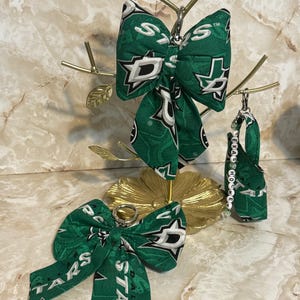 May include: Green and white Dallas Stars themed fabric bows and a small bag, displayed on a gold-colored decorative stand. The bows feature the team's logo and the word "STARS". A small gold bird sits atop the stand.