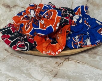 May include: A collection of fabric hair scrunchies featuring various sports team logos and colors, including orange, blue, black, and white patterns. The scrunchies are piled together in a wooden bowl, displaying multiple team-themed prints.