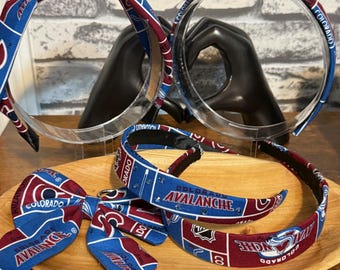 May include: Headbands and a bow tie featuring a blue and burgundy pattern with the text "COLORADO AVALANCHE". The headbands have a clear plastic base. The bow tie is also made of the same patterned fabric. The items are displayed on a wooden tray.