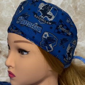 May include: A blue scrub cap featuring a repeating pattern of the Ravenclaw house crest, eagle emblems, and the word Ravenclaw in white and black lettering. The fabric has a checkered background detail and small star accents throughout the design.