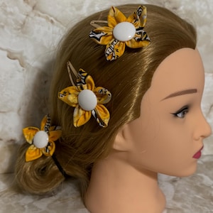May include: Three yellow and black floral hair clips with white centers are displayed on a mannequin head with blonde hair. The clips are attached to the hair with gold-colored metal clips. The flowers have a patterned design.