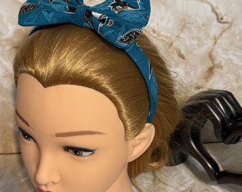 Pode incluir: Tiara de tecido azul-petróleo com um laço grande. O laço e a tiara são estampados com um design com gráficos pretos e laranja. A tiara é exibida em uma cabeça de manequim com cabelo loiro.