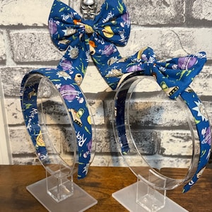 May include: Two blue headbands with matching bow ties. The headbands and bows are decorated with a cartoon design featuring characters and elements from the Rick and Morty animated series. The headbands are displayed on clear plastic stands.