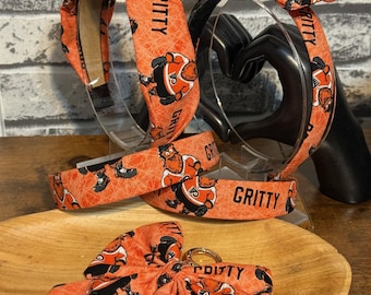 May include: A collection of orange headbands, bow ties, and wristbands with a repeating mascot graphic and the word "GRITTY". The accessories are displayed on clear stands and a wooden tray. The mascot is wearing a hockey uniform.