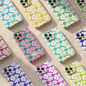 May include: A collection of floral-patterned phone cases in various colors, including blue, green, pink, and purple. Each case features a different color flower design and a two-letter initial. The cases are arranged on a light-colored surface.