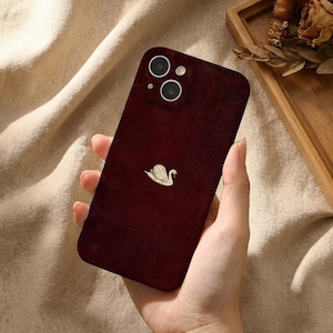 Puede incluir: Una funda para smartphone de color burdeos intenso con un sutil patrón floral oscuro y una pequeña ilustración de un cisne blanco centrada. La funda incluye un recorte para la cámara dual en la parte superior izquierda y un acabado liso.