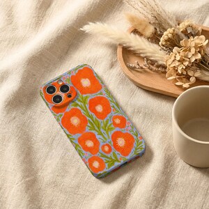 Puede incluir: Una funda rectangular para smartphone con un patr&oacute;n vibrante de flores de amapola de color naranja brillante con centros amarillos y tallos verdes sobre un fondo azul b&iacute;garo claro. La funda incluye un recorte para una c&aacute;mara triple.