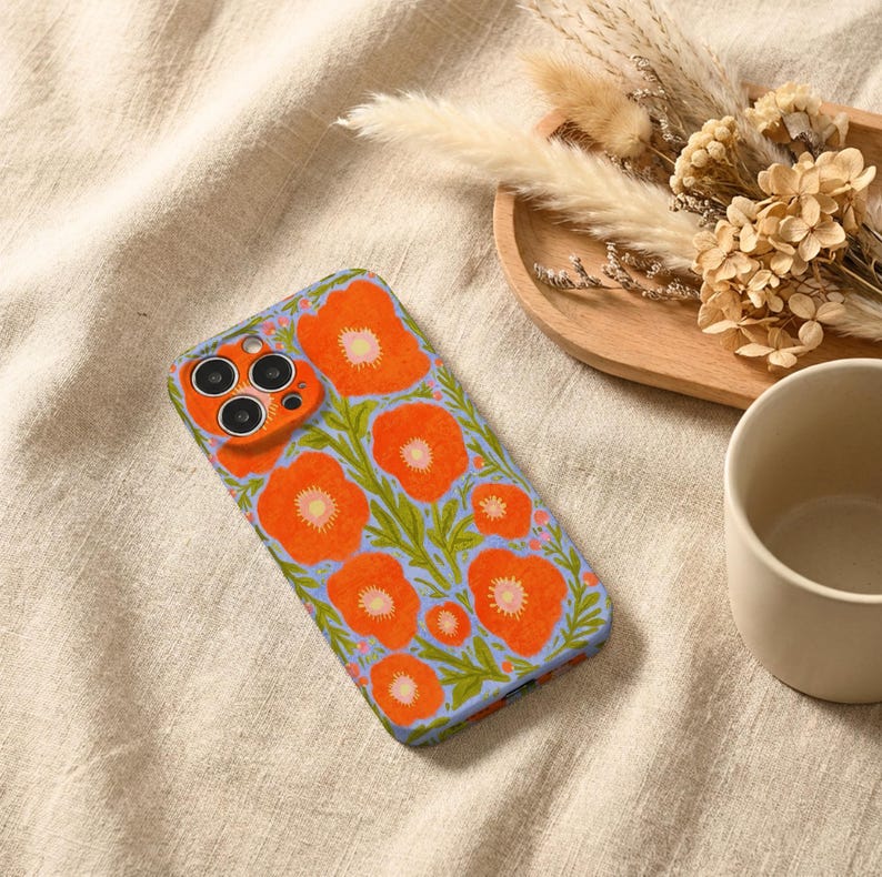 Puede incluir: Una funda rectangular para smartphone con un patr&oacute;n vibrante de flores de amapola de color naranja brillante con centros amarillos y tallos verdes sobre un fondo azul b&iacute;garo claro. La funda incluye un recorte para una c&aacute;mara triple.