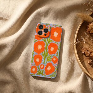 Puede incluir: Una funda rectangular para tel&eacute;fono con un vibrante estampado floral de amapolas de color naranja brillante y follaje verde sobre un fondo azul b&iacute;garo claro. La funda incluye una secci&oacute;n elevada recortada para la c&aacute;mara en la esquina superior.