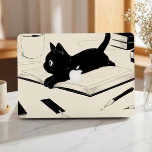 Könnte beinhalten: Laptop-Hülle mit einer Illustration einer schwarzen Katze. Die Katze liegt auf einem offenen Buch, mit einem Apple-Logo. Das Design umfasst eine Kaffeetasse, ein Tintenfass und einen Stift, alles in Schwarz und Weiß.