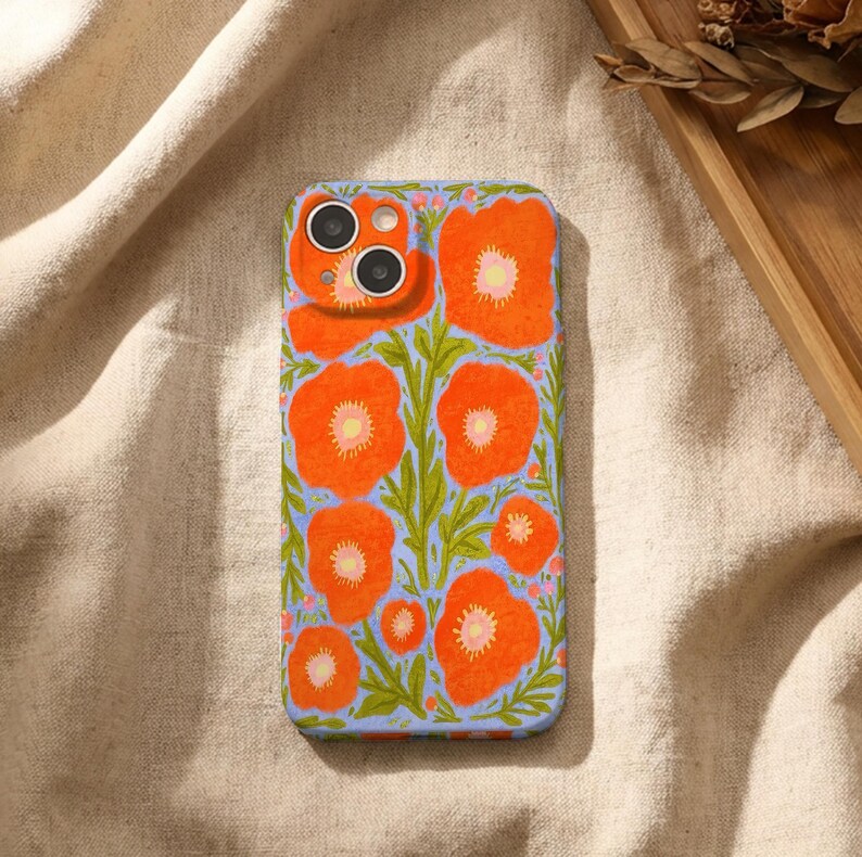 Puede incluir: Una funda rectangular para smartphone con una ilustraci&oacute;n vibrante de amapolas de color naranja brillante con centros amarillos y tallos verdes sobre un fondo azul b&iacute;garo claro. La funda tiene un recorte para un sistema de c&aacute;mara dual.