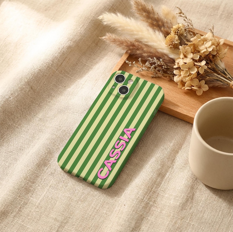 Puede incluir: Una funda de tel&eacute;fono a rayas verdes y verde claro con el nombre "CASSIA" en letras rosas. La funda est&aacute; sobre una superficie beige con flores secas y una taza beige en el fondo.