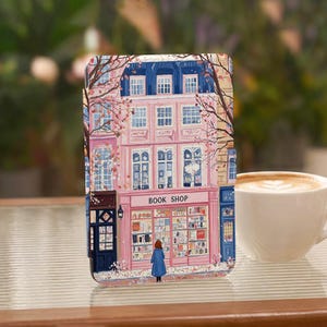 May include: A rectangular e-reader case featuring a colorful illustration of a pink building with a book shop storefront. A person in a blue coat stands in front of the shop window. The design includes bare tree branches and blue roof details.