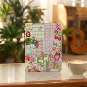 Könnte beinhalten: Eine rechteckige Tablethülle mit einem bunten Patchwork-Design in grünen und rosafarbenen Karomustern. Die Oberfläche ist mit verschiedenen Illustrationen verziert, darunter eine grüne Raupe, Erdbeeren, Feigen, Blumen und ein gelber Stern.